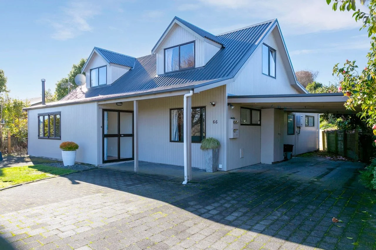 Front view of a modern two-story house with a pitched metal roof, white siding, large windows, and a covered carport to the right. The house number 66 is visible near the entrance, with potted plants and a driveway in the foreground.