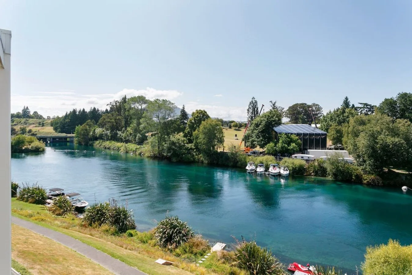 A scenic view of a river with clear blue water, surrounded by lush green trees and a grassy area with parked boats and cars. There is a large covered outdoor stage or tent structure on the right and a bridge in the distance.