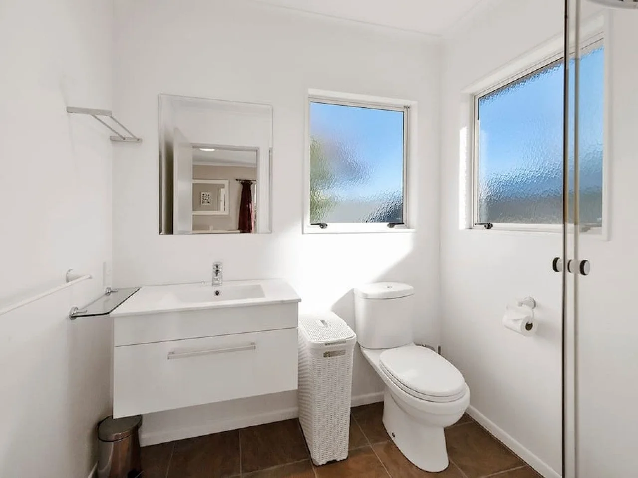A modern, minimalist bathroom with white walls, a white vanity with sink, a mirror, a toilet, two frosted windows, a laundry basket, and a small closet with black handles.