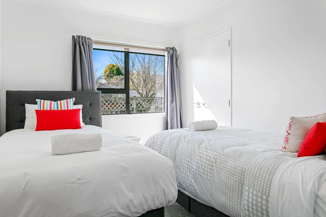 A bedroom with two beds, one with a dark gray headboard and colorful striped pillows, the other with white bedding and red pillows, a window with gray curtains showing trees outside, and a closed white door.