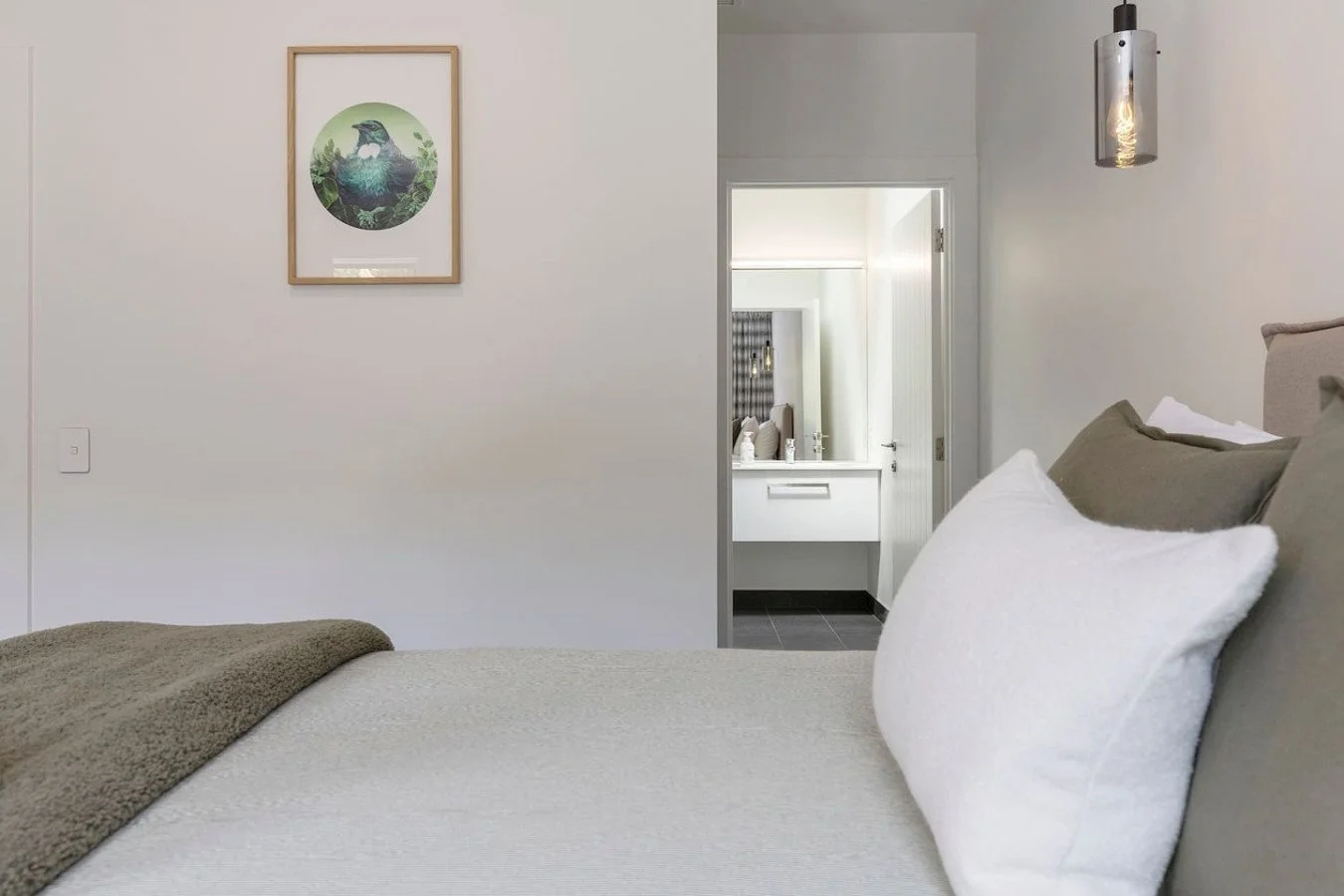 Hotel room with neatly made bed, white and beige pillows, and an open door to a bathroom with a white sink and countertop, hanging light fixture, and minimalist decor.