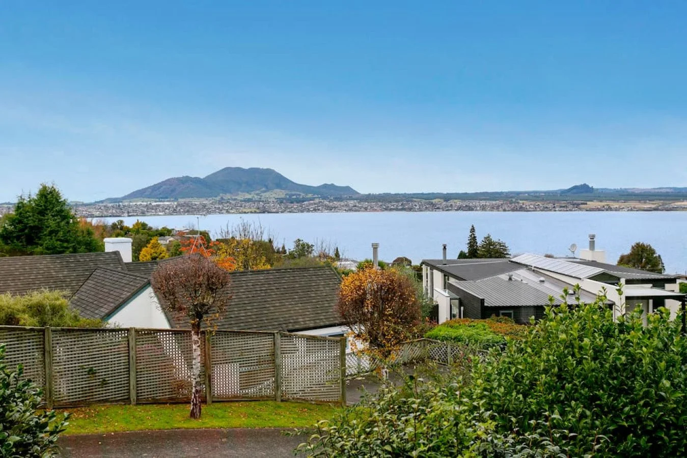 View of a lake with houses and trees in the foreground, mountains in the background, under a clear blue sky.