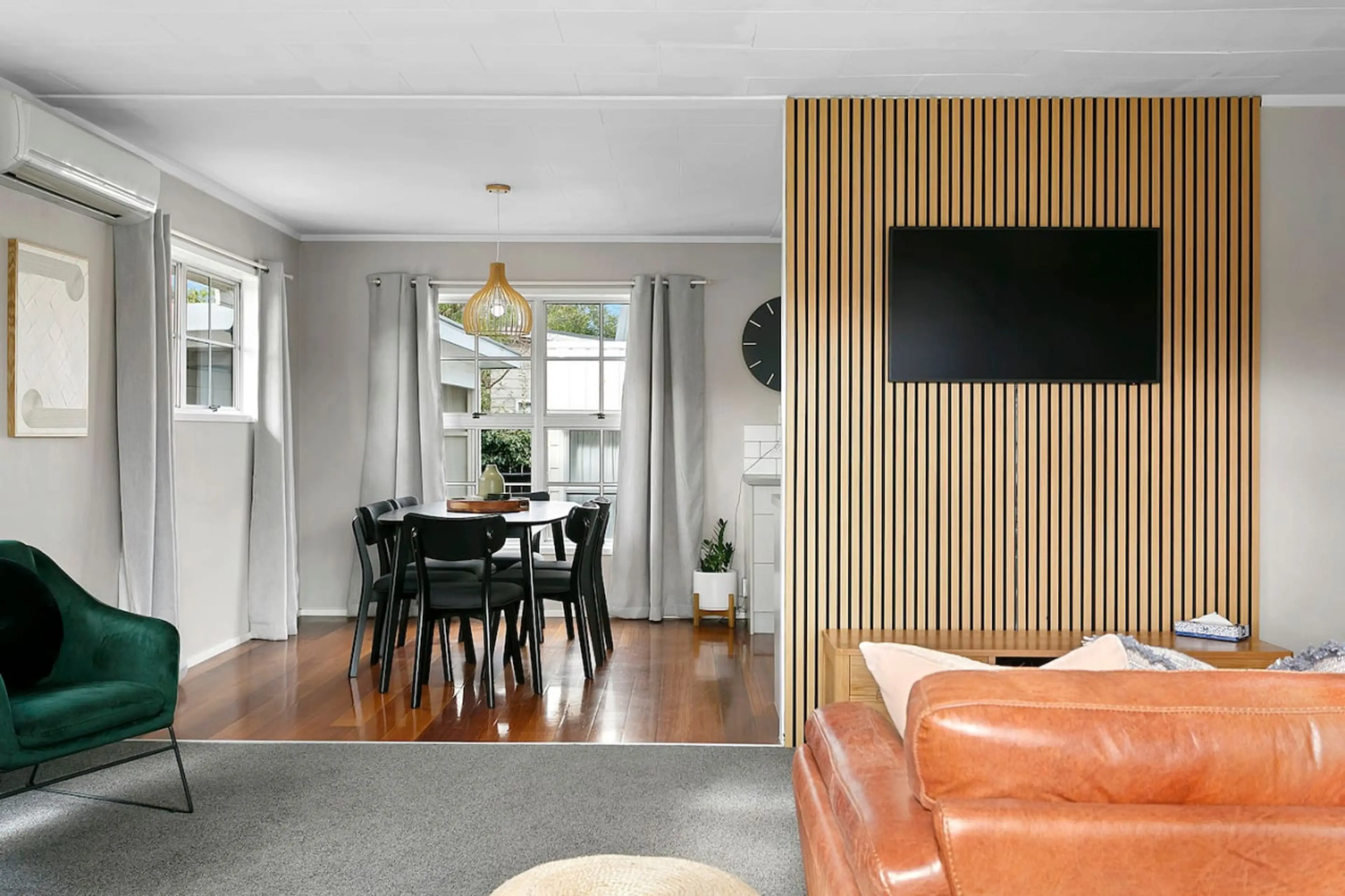 Interior view of a living and dining area with wooden floor, green armchair, black dining chairs around a table, a wall-mounted TV on a slatted wooden divider, and large windows with white curtains.