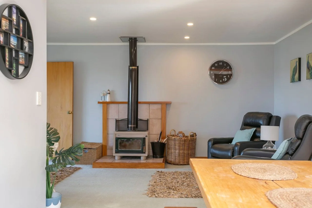 Living room with a wood stove, a wicker basket, black leather recliners, a side table with a lamp, wall art, a wall clock, and a potted plant.