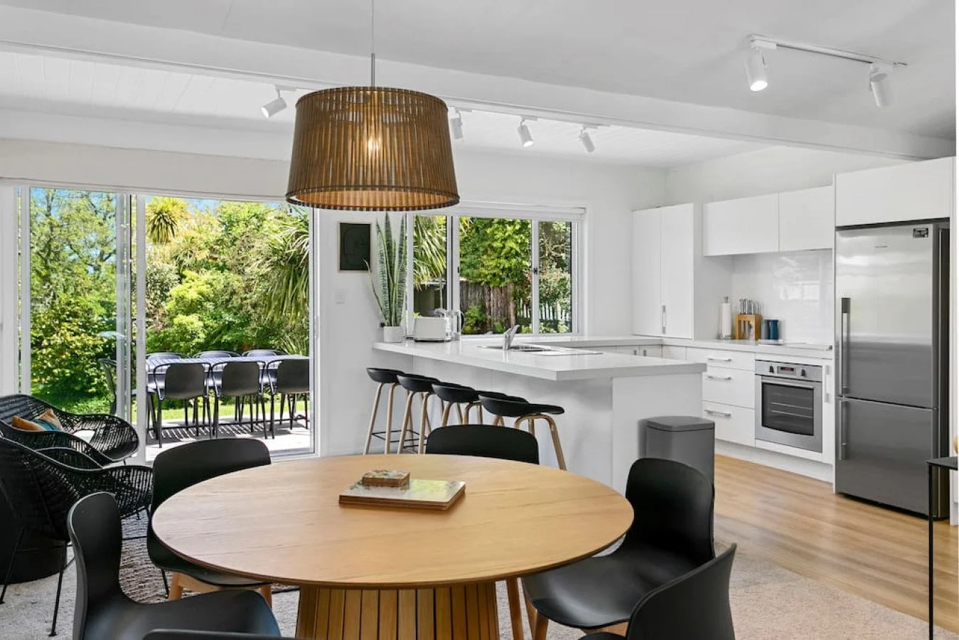 Modern white kitchen with a dining area, black chairs around a wooden table, stainless steel appliances, and large windows showing greenery outside.