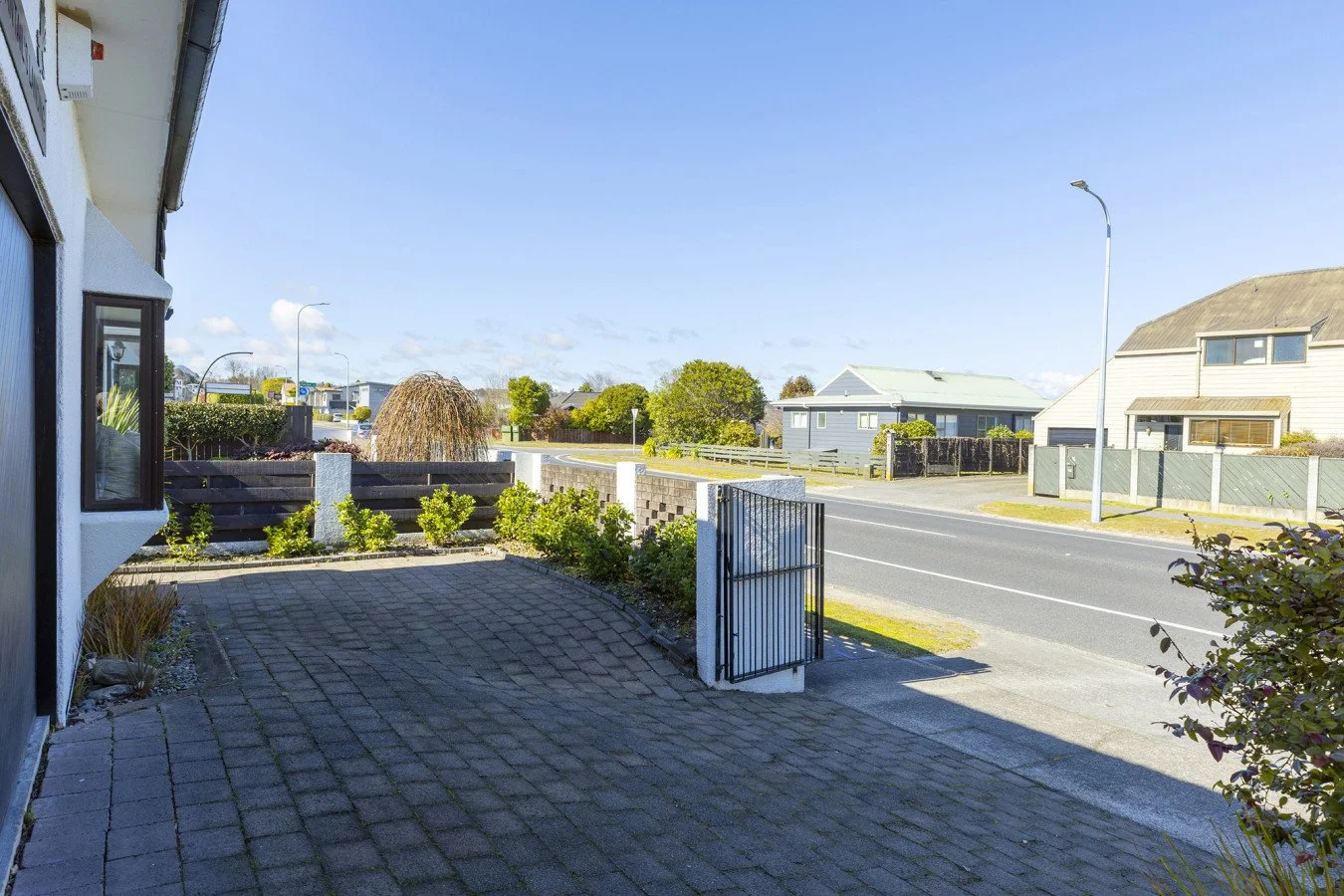 A suburban street viewed from a driveway with brick paving, showing a road with a lamp post, residential houses, and a clear blue sky.