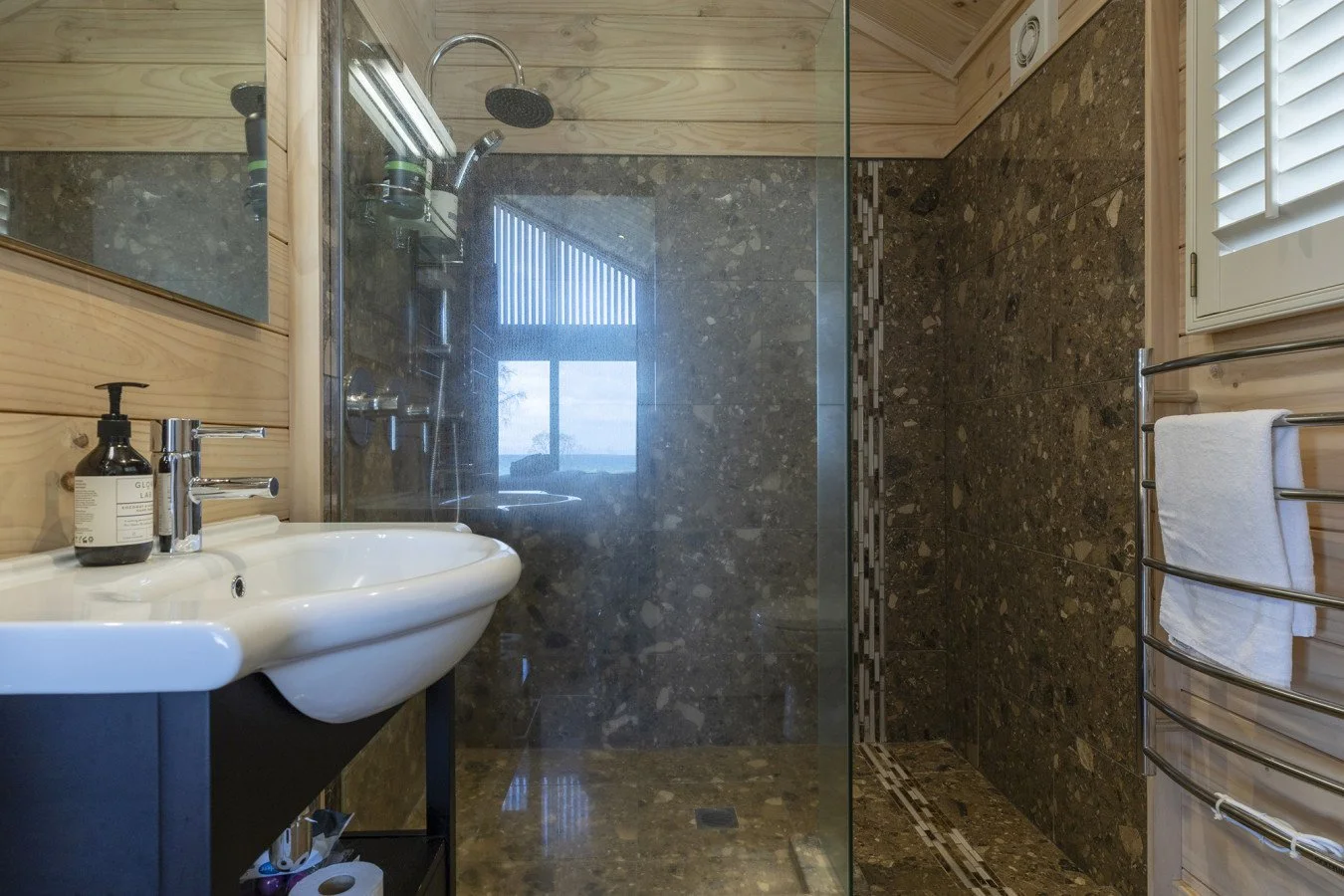 Modern bathroom with a white sink, black shelving, brown marble tiles on the wall and floor, a glass shower enclosure with rainfall shower head, a window with white shutters, and a towel rack with a white towel.