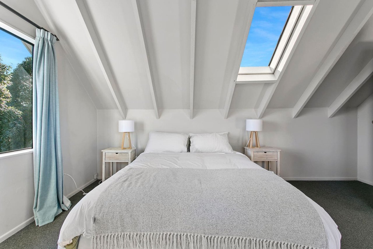 A bedroom with a sloped ceiling, a skylight window, a large bed with white bedding, two nightstands with lamps, a window with blue curtains, and trees outside.