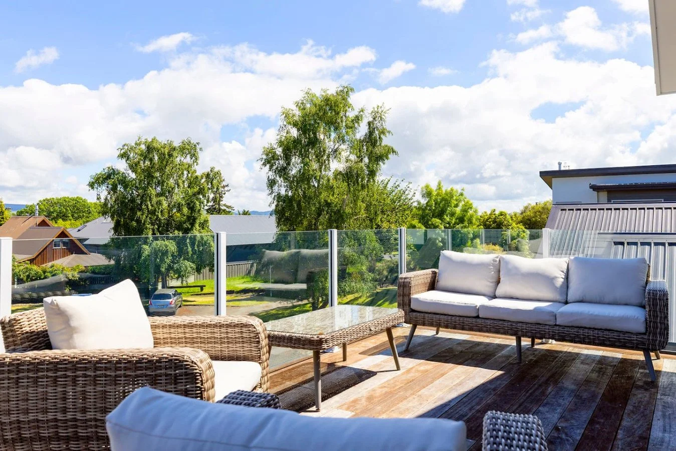 Outdoor patio with wicker furniture and white cushions, glass railing, wooden deck, green trees in the background, partly cloudy sky.
