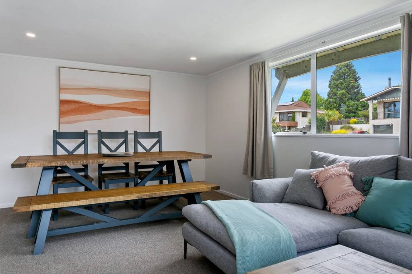 Living room with a gray sofa, decorative pillows, and a wooden dining table with matching benches and chairs, large window with curtains, and framed artwork on white wall.