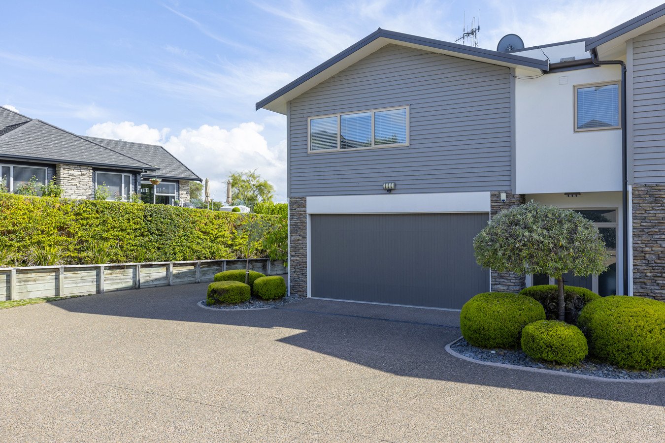 Modern two-story house with gray siding, stone accents, and an attached garage, surrounded by neatly trimmed bushes and landscaping, under a partly cloudy sky.