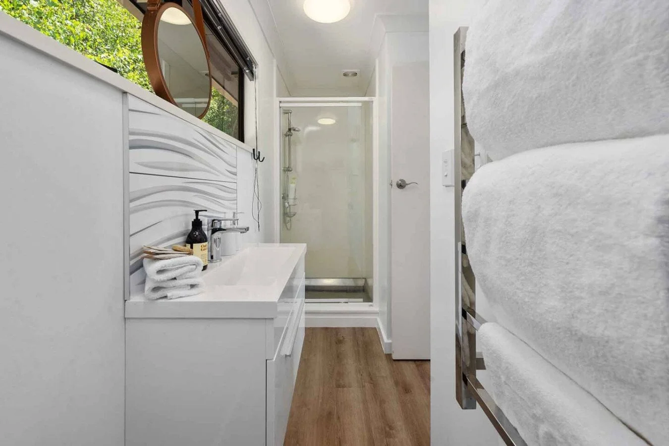 A modern, compact bathroom with a white vanity, black soap dispenser, and folded towels, a shower stall with glass doors, and a nearby stack of white towels on a metal rack.