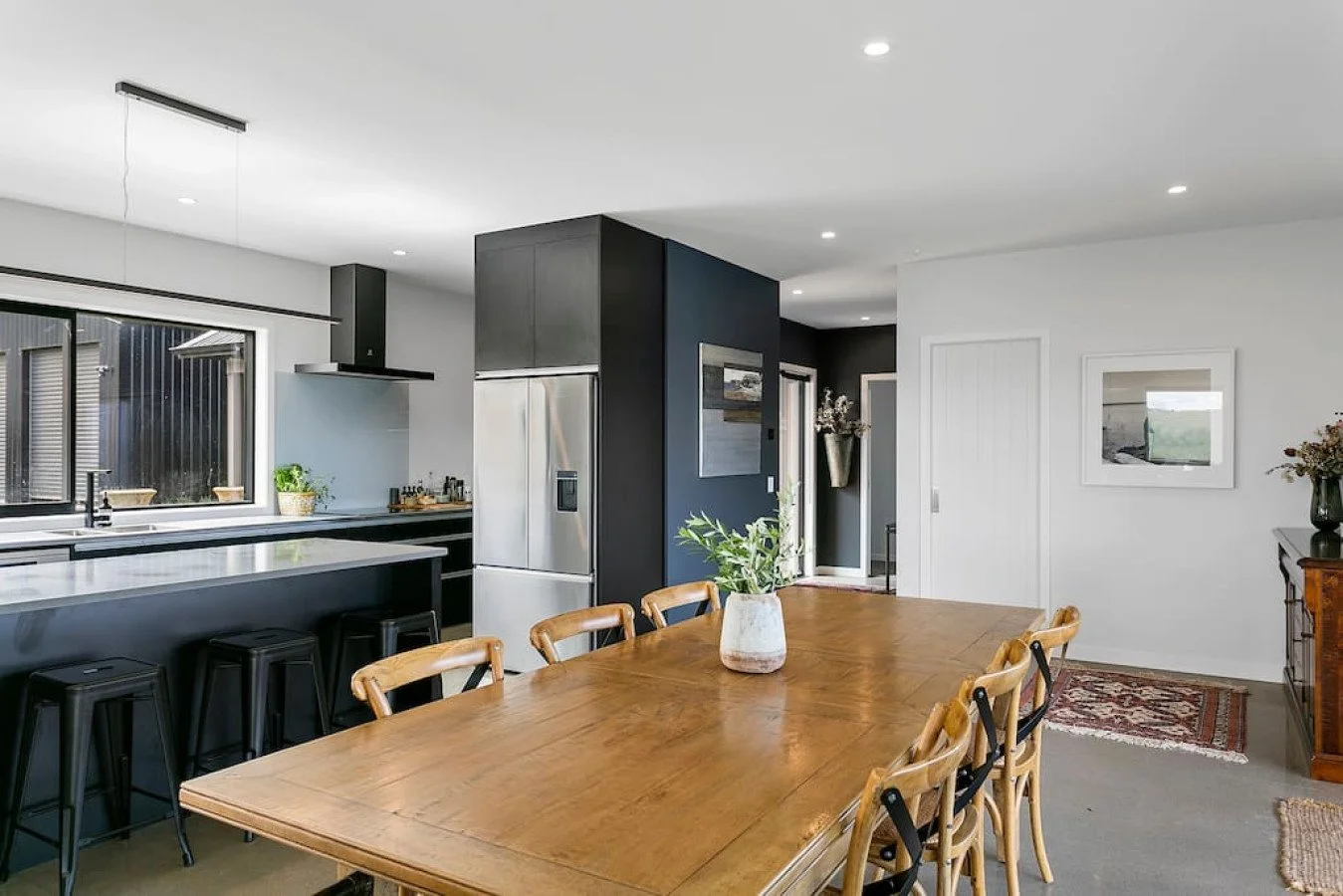 Open-concept kitchen and dining area with a wooden dining table, beige chairs, black kitchen island with bar stools, gray and white walls, stainless steel refrigerator, black cabinetry, and decorative plants and artwork.