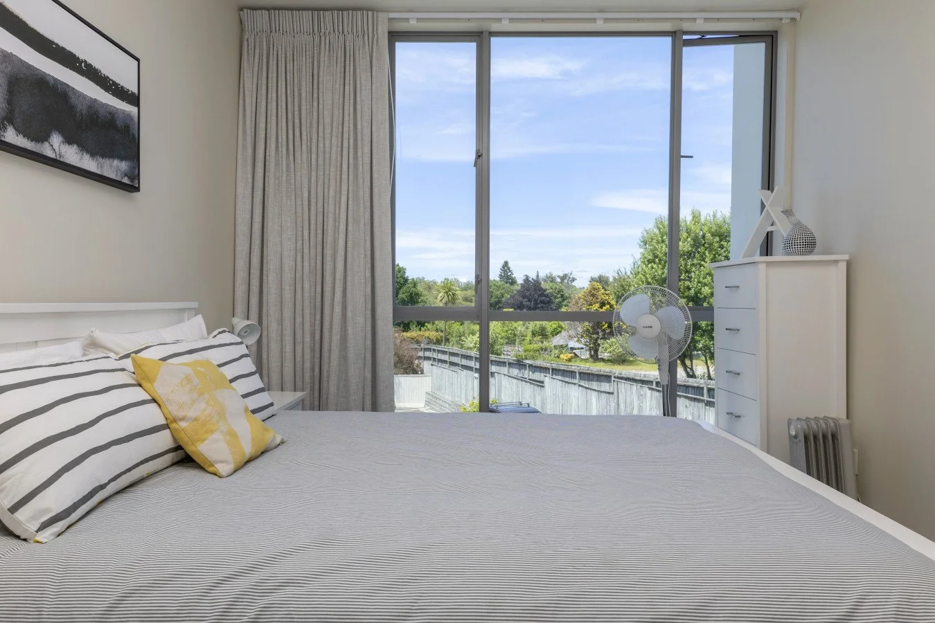 Bright bedroom with large window showing trees and sky, bed with striped pillows and yellow accent pillow, white dresser with decorative items, standing fan, and abstract wall art.