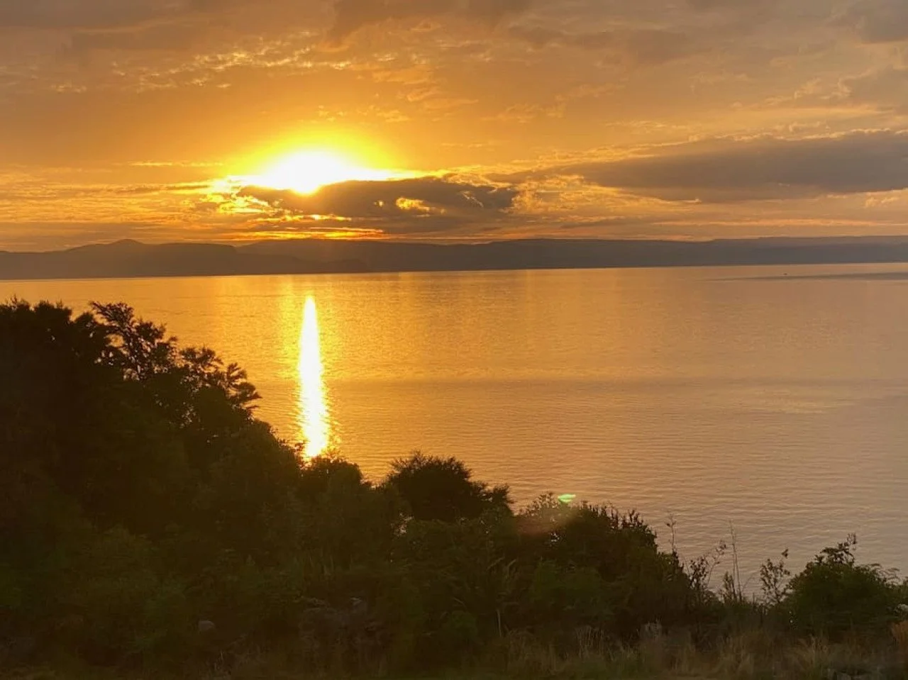 Sun setting over a large body of water with trees in the foreground, clouds in the sky, and the sun's reflection on the water.