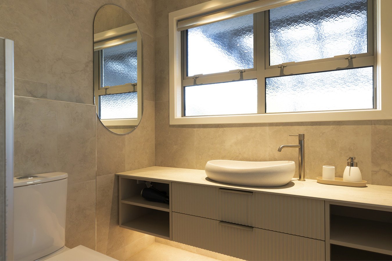 Modern bathroom with a beige vanity, vessel sink, towel, soap dispenser, and a window with textured glass.