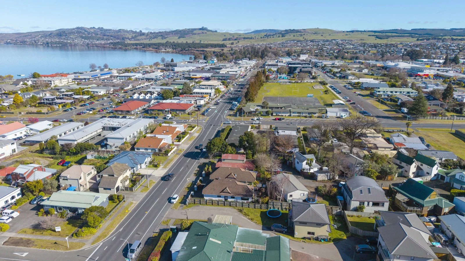 Aerial view of a suburban neighborhood near a lake, showing residential houses, roads, and a commercial area with parking lots, surrounded by green hills under a partly cloudy sky.