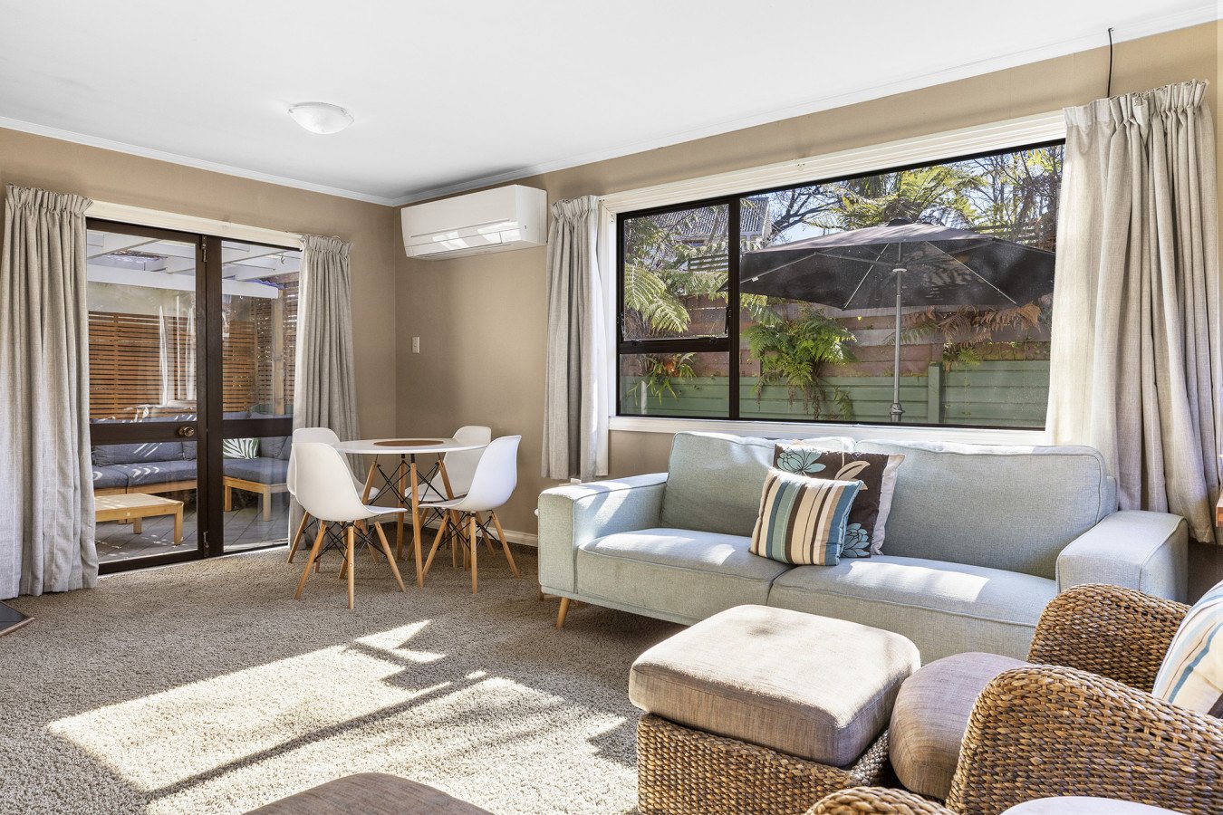 Living room with a beige carpet, a light green sofa with striped and floral pillows, a rattan armchair, a small round dining table with four white chairs, large windows and sliding glass doors with curtains, and an outdoor patio with a table, patio c