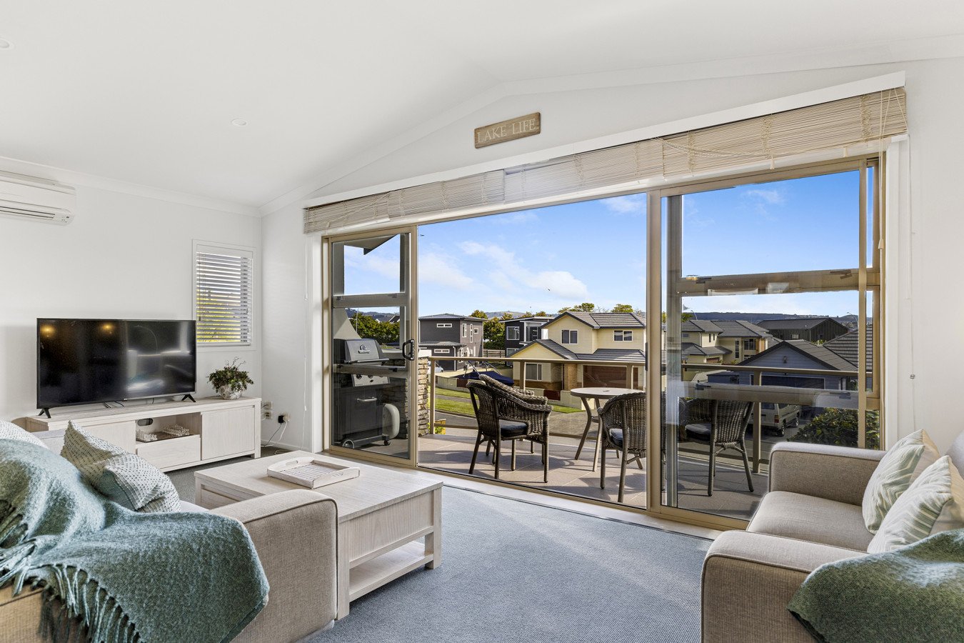 Living room with large sliding glass doors leading to a balcony with outdoor furniture, overlooking a neighborhood with houses under a clear blue sky.