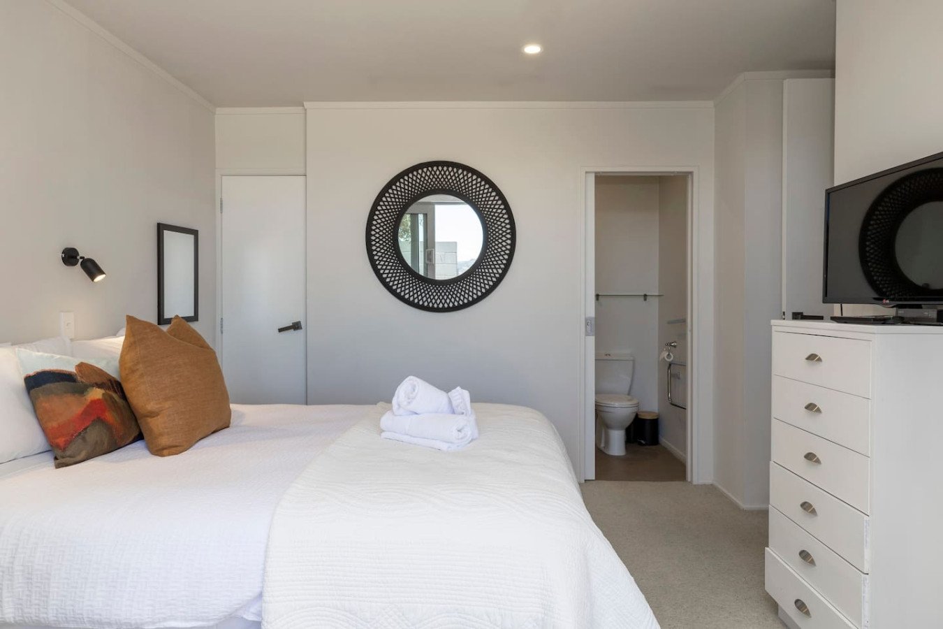 A bedroom with a white bed, pillows, and towels, a round mirror on the wall, and a small bathroom visible through an open door.