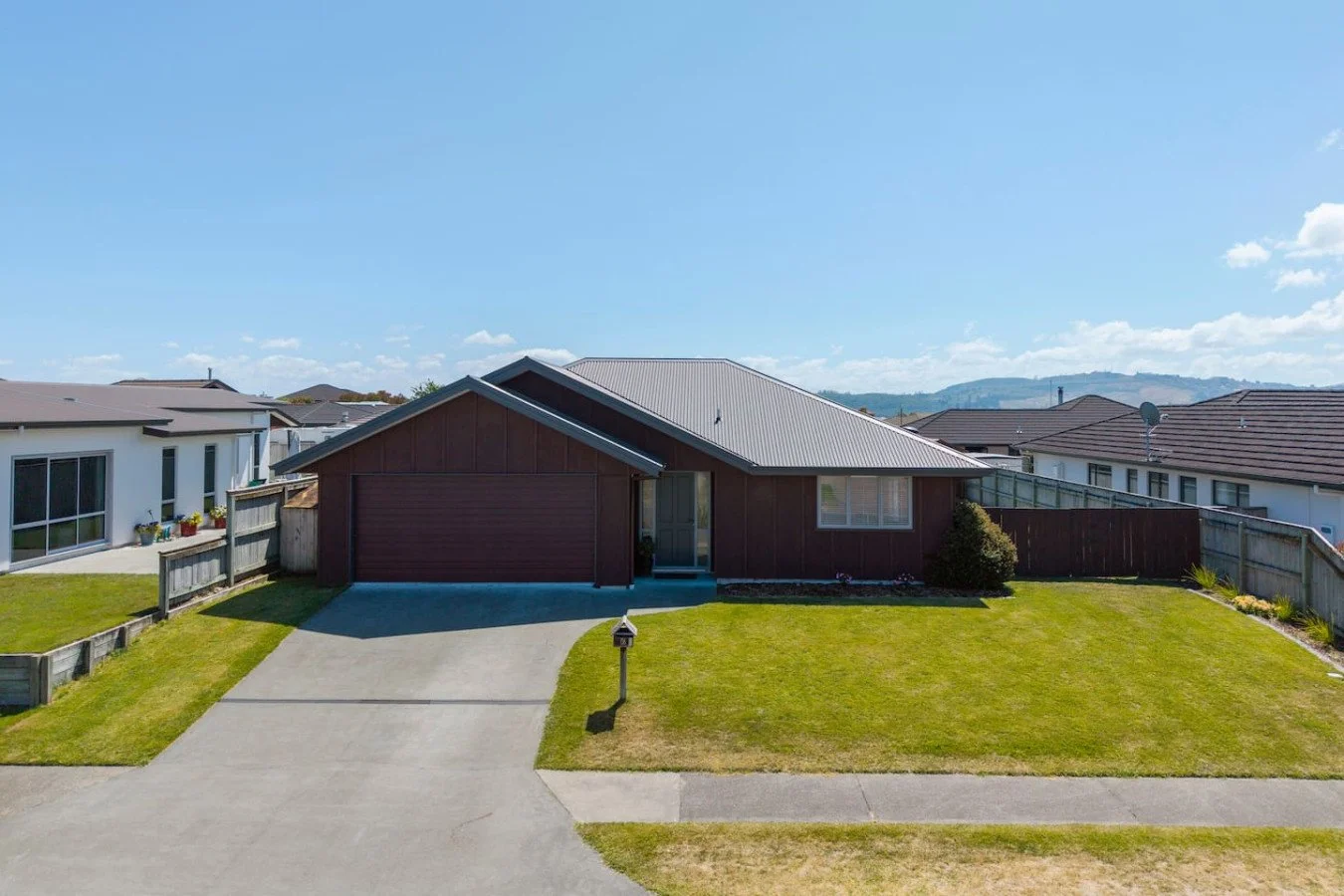 Front view of a single-story house with a brown exterior, gray metal roof, and a driveway leading to a two-car garage. The house has a small front lawn with a mailbox near the sidewalk, and neighboring houses are visible on either side. Mountains are