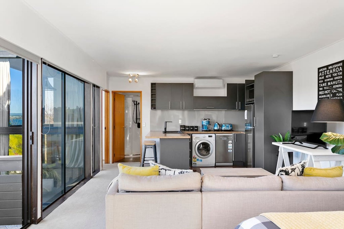 Open-plan living room and kitchen with sliding glass doors, beige sofa with yellow pillows, gray kitchen cabinets, and modern appliances.