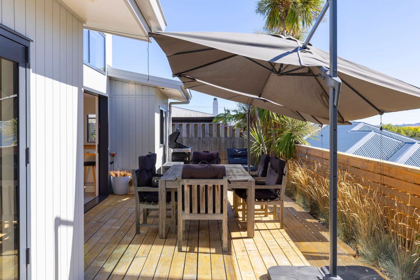 Outdoor patio with wooden deck, large umbrella, dining table, and chairs, surrounded by a wooden fence and plants, with part of a house and neighboring rooftops visible.