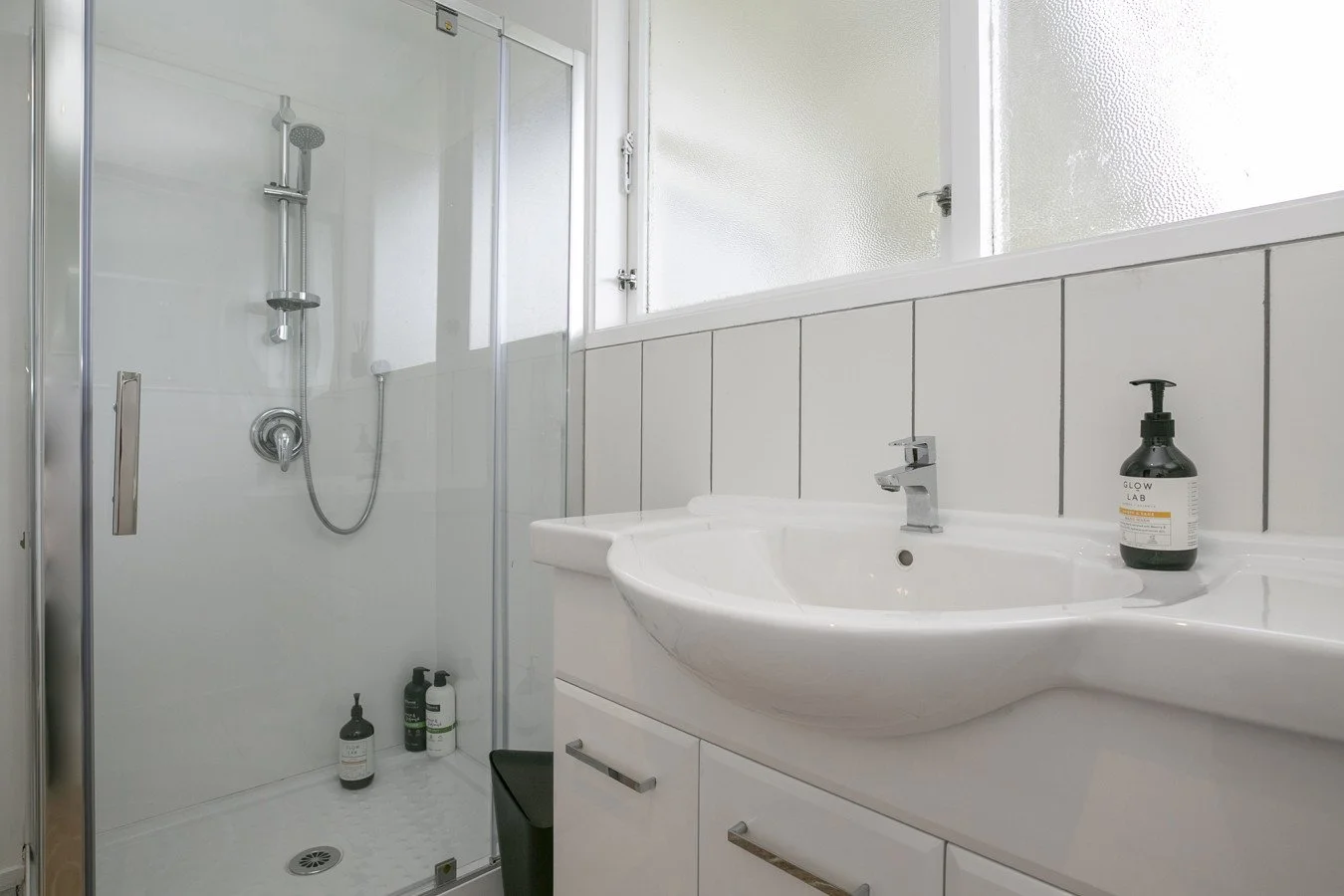 A bathroom with a glass shower enclosure on the left and a white sink with a chrome faucet on the right. There are black liquid soap bottles on the sink and a black waste bin below. The wall behind the sink is tiled with white panels, and a frosted w