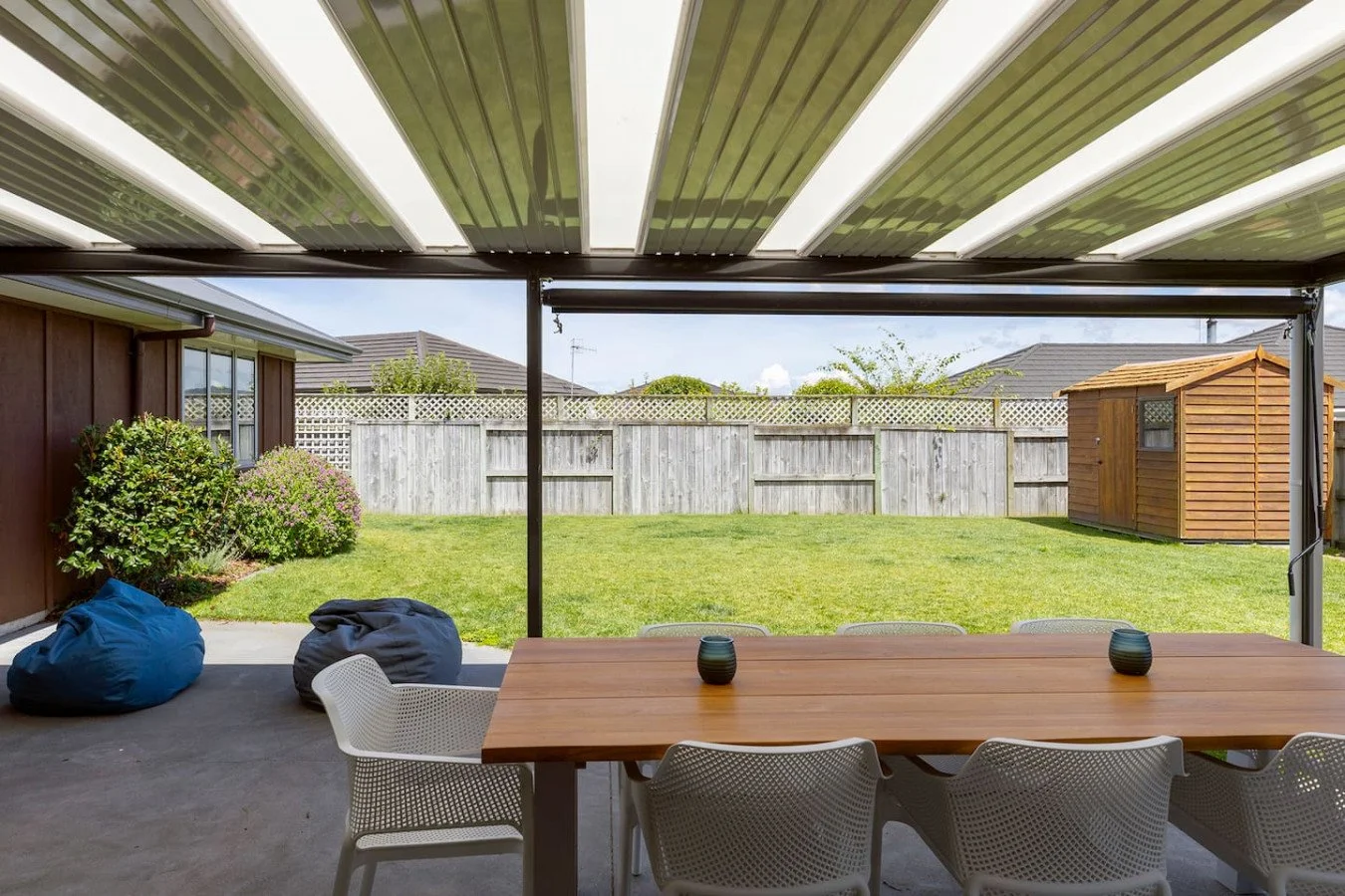 Covered backyard patio with a wooden table, white chairs, and decorative vases, overlooking a grassy yard with a wooden fence and garden shed.