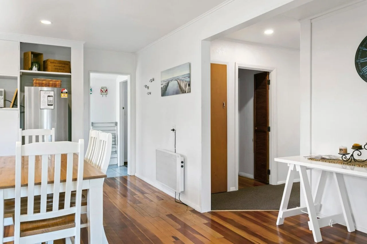 Interior view of a home with white walls, wooden floors, a dining table with white chairs, a small white table with candles, and a hallway leading to other rooms.