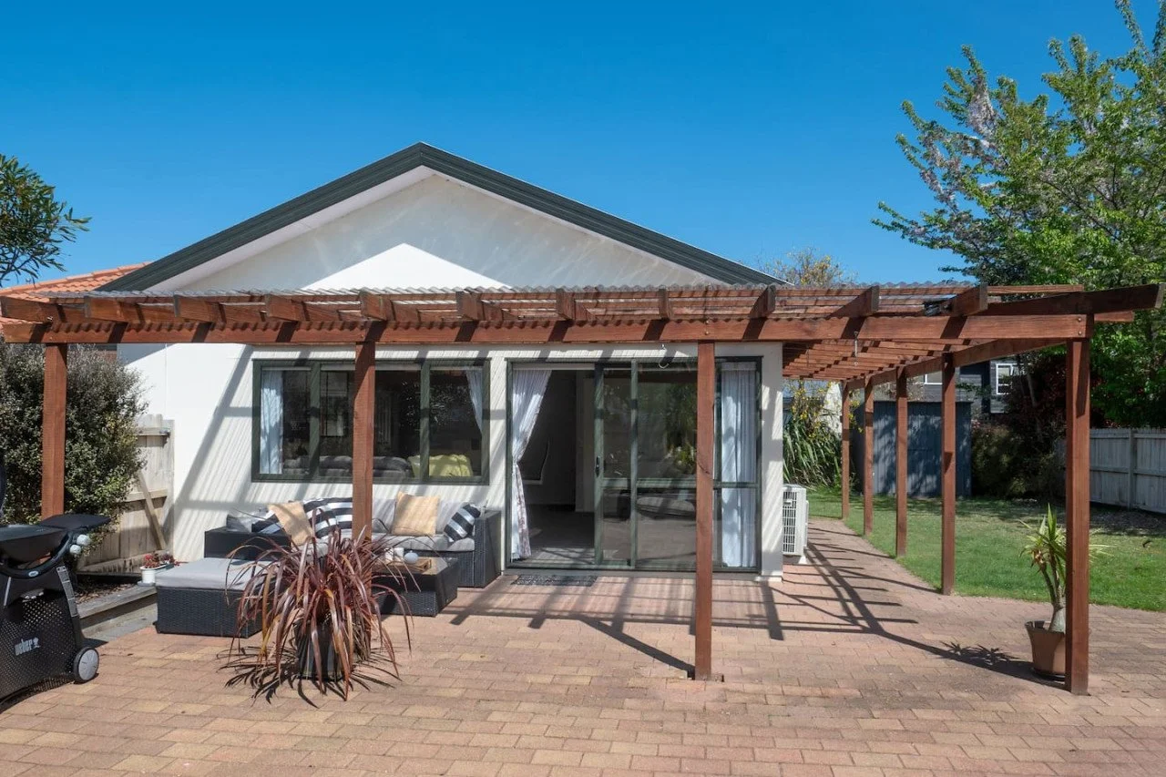 Backyard patio with wooden pergola covering a sitting area with a black outdoor couch and a potted plant, overlooking a white house with sliding glass doors and greenery, under a clear blue sky.