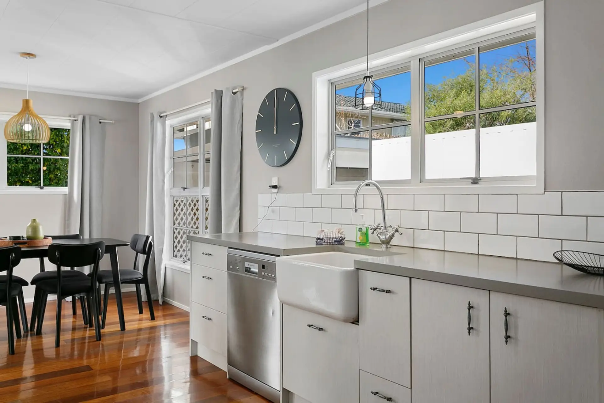 Modern kitchen with white cabinets, farmhouse sink, large window, and hardwood floors, with a dining area featuring black chairs and a wooden table.