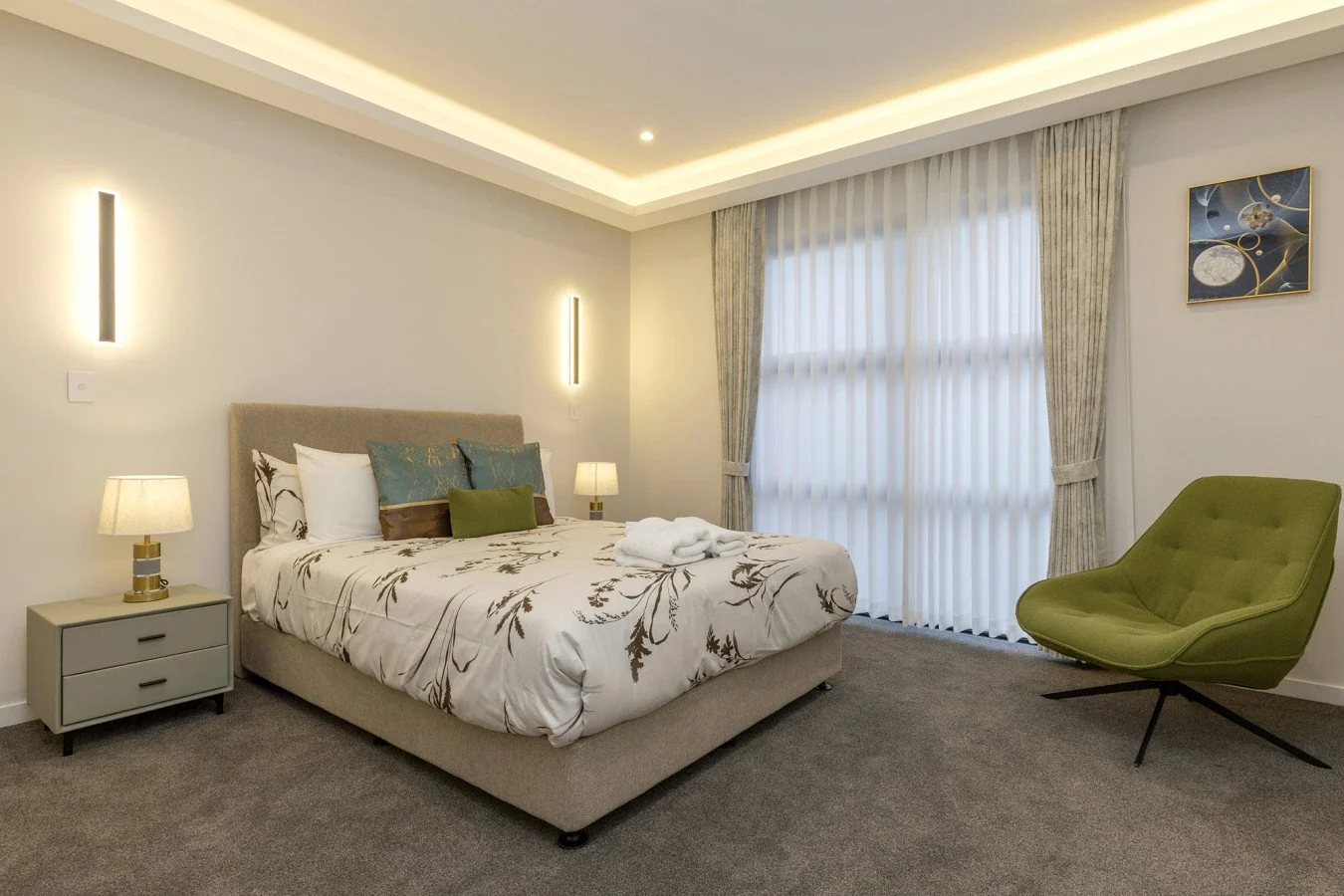 Modern hotel bedroom with a bed, nightstand, lamps, green lounge chair, and large window with curtains.