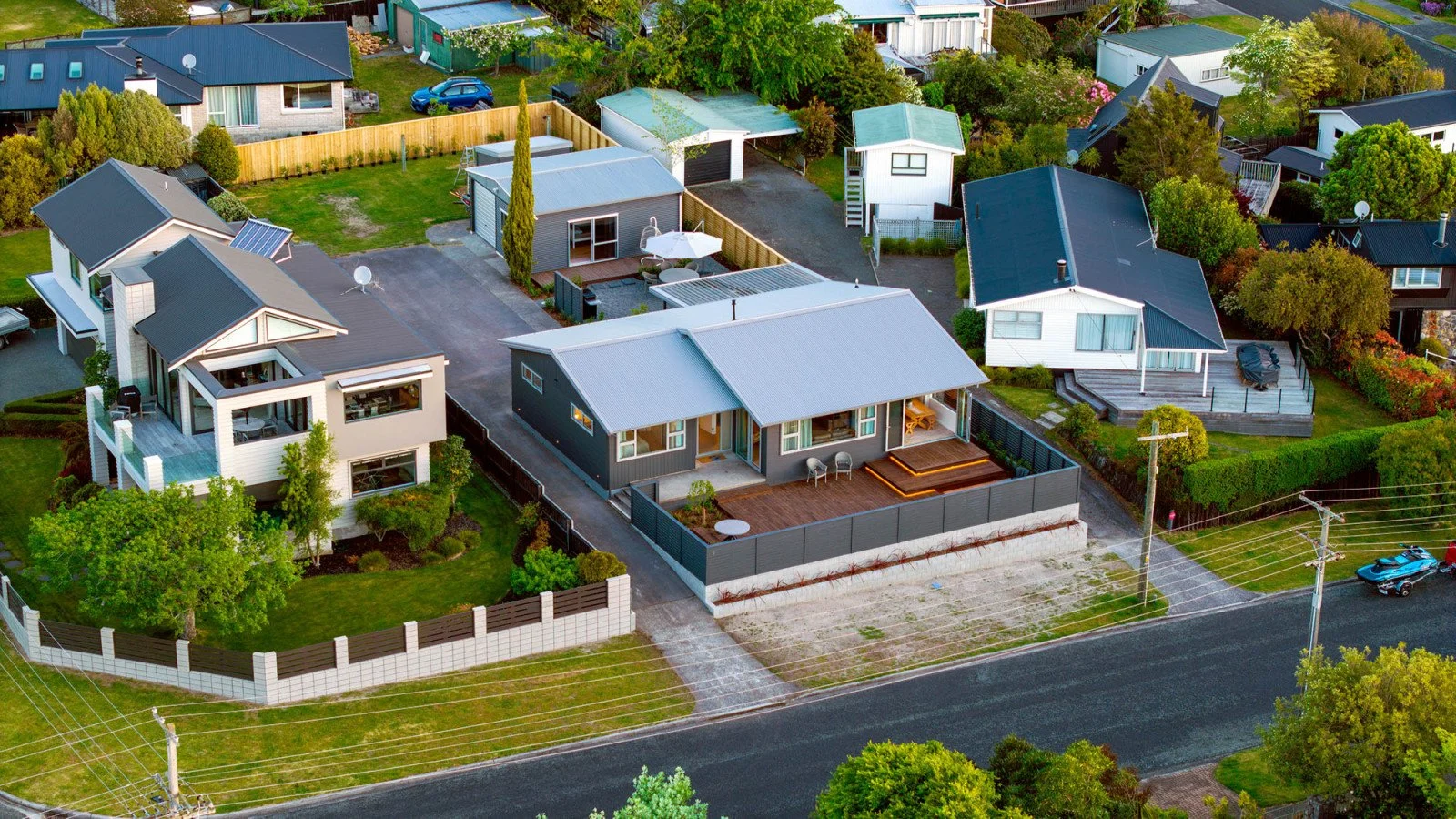 Aerial view of a suburban neighborhood with several modern houses, green lawns, trees, driveways, and fenced yards. There are cars parked on the street and in driveways, with a mix of one and two-story homes with different roof styles and colors.