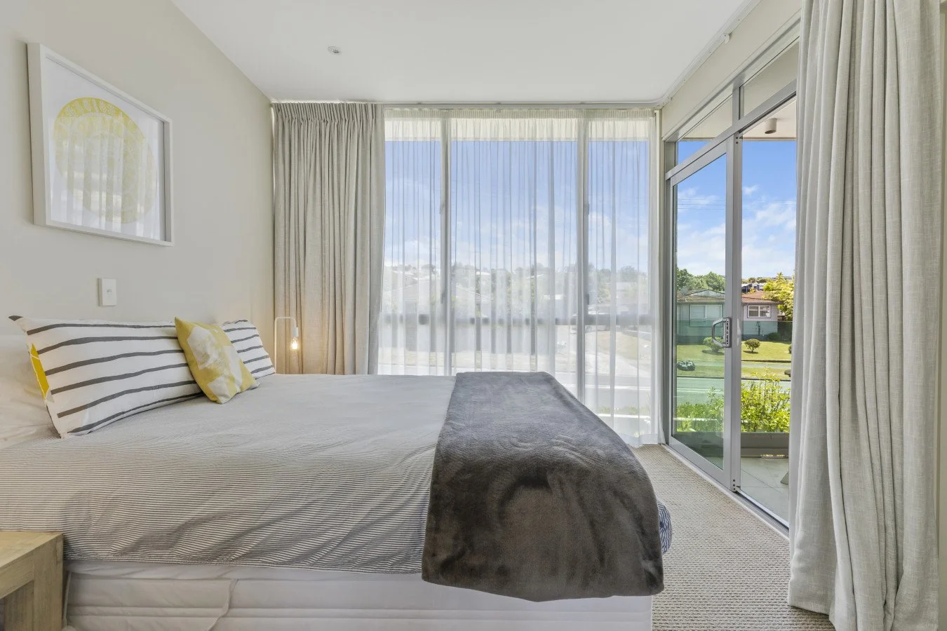 Bright bedroom with large windows covered by sheer curtains, a bed with striped and yellow pillows, and a small bedside table.