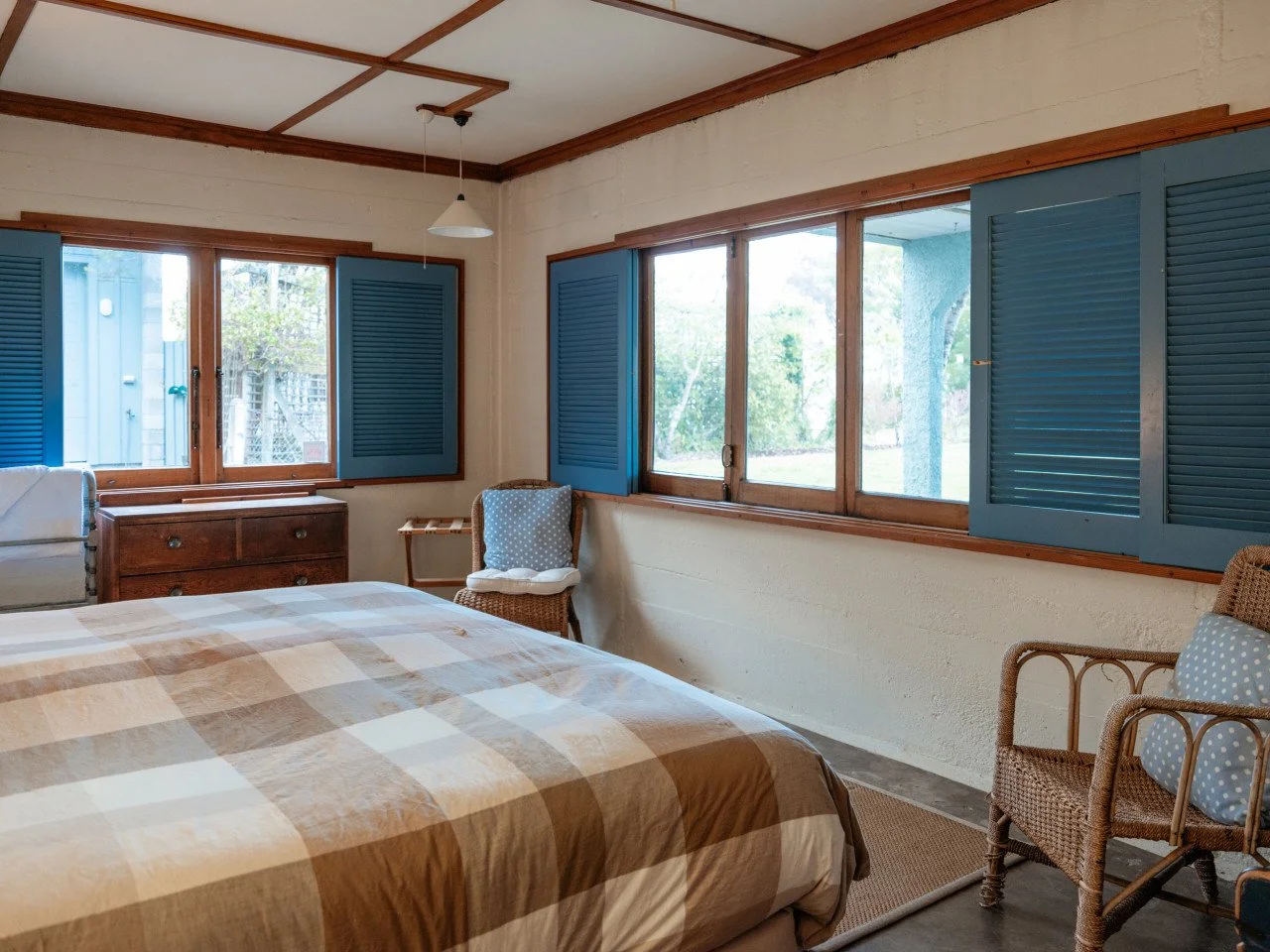 A cozy bedroom with a bed covered in a checkered beige and brown blanket, surrounded by wicker chairs with blue polka-dotted cushions, and large windows with blue shutters showing a green outdoor scene.