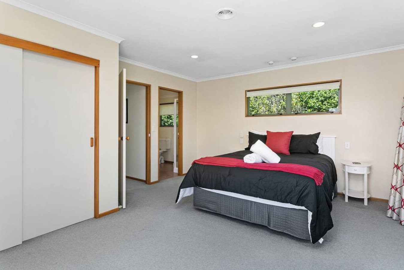 Bedroom with a bed covered in black and red bedding, white rolled towel, black and red pillows, window with greenery outside, and a small white side table.