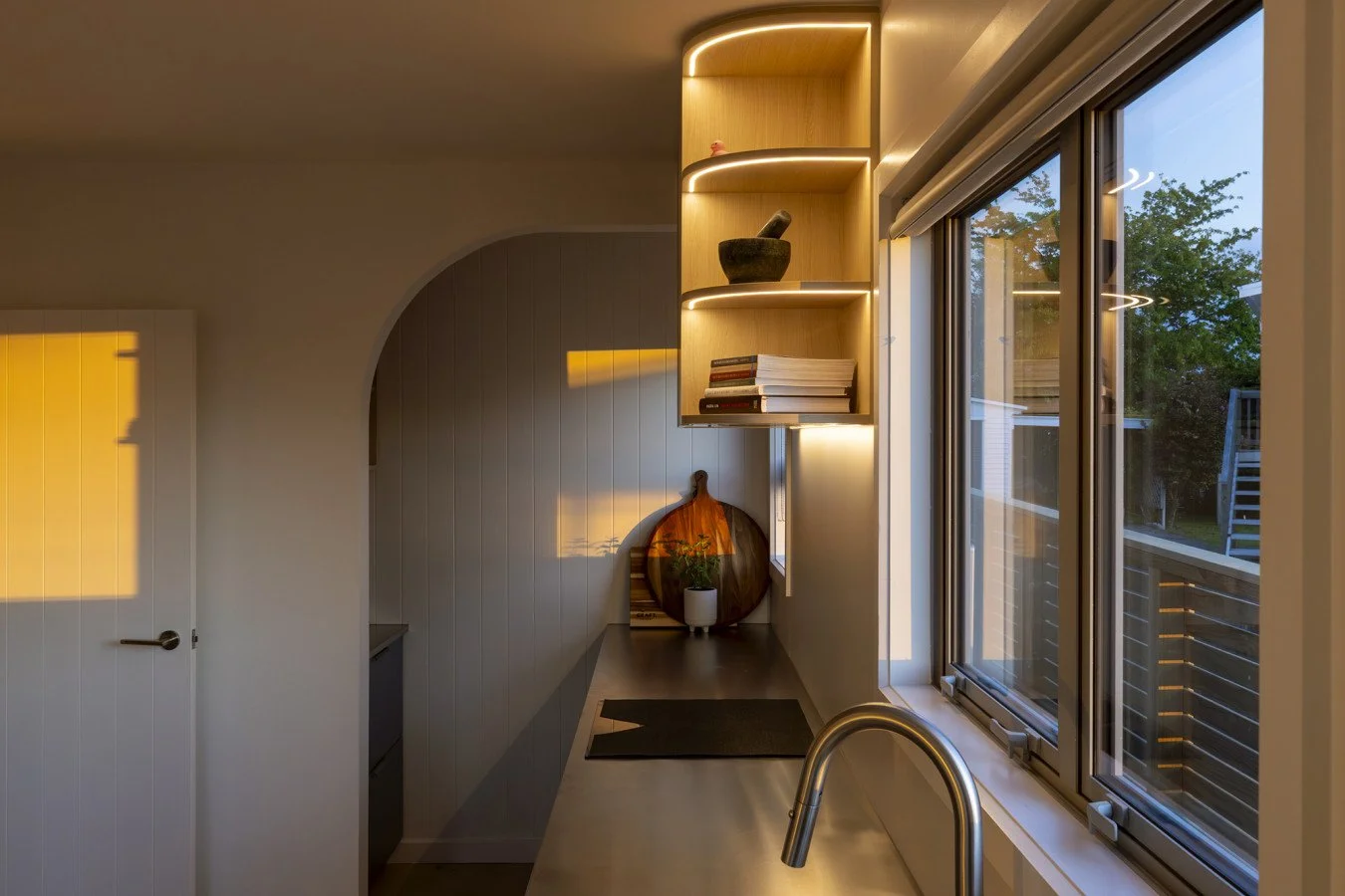 Modern kitchen with a window showing outdoor greenery, open shelves with books and decorative items, and a wooden cutting board with a plant on it.