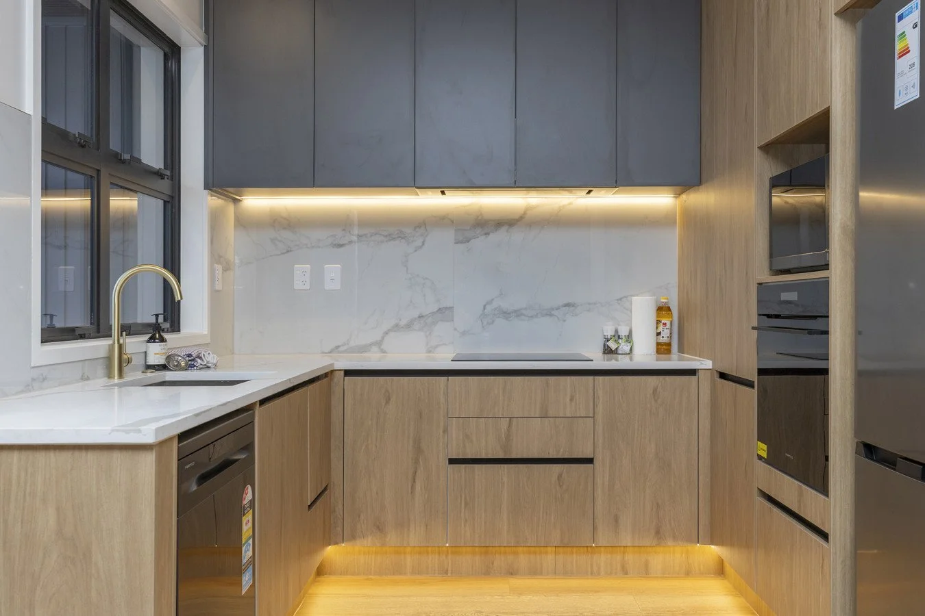 Modern kitchen with wood and gray cabinetry, marble backsplash, LED under-cabinet lighting, stainless steel appliances, and a black induction cooktop.