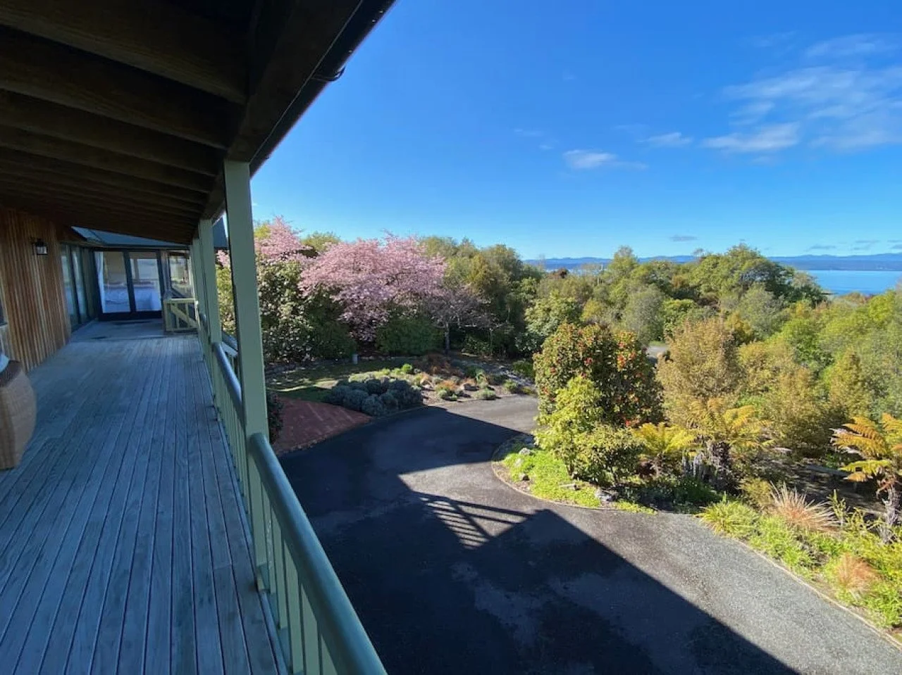 View from a balcony overlooking a garden with flowering trees, a driveway, and a distant body of water with hills on the horizon under a blue sky.