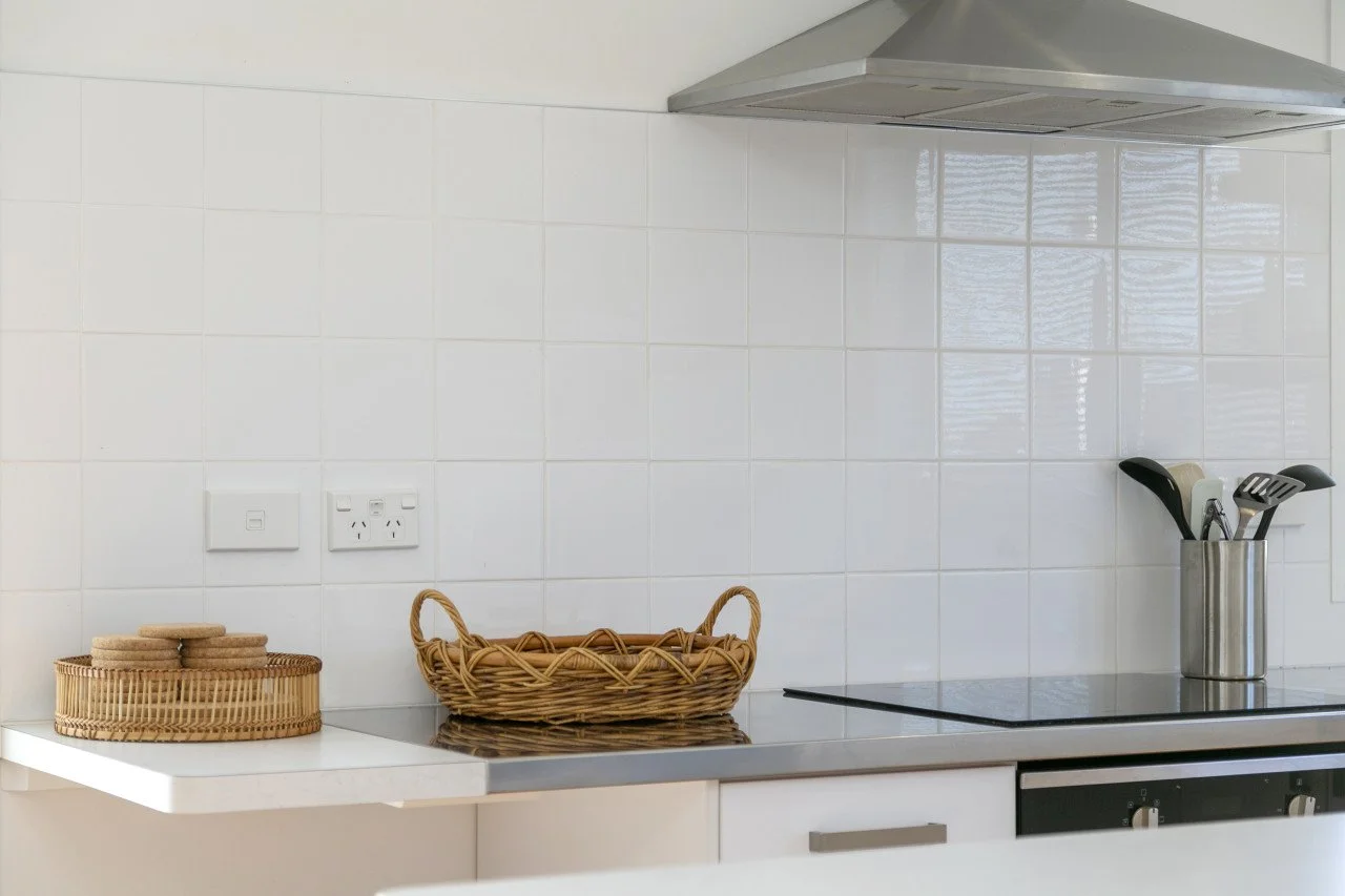 A modern kitchen countertop with a round wicker basket, a rectangular wicker tray with cookies, and a stainless steel utensil holder with spatulas and spoons.