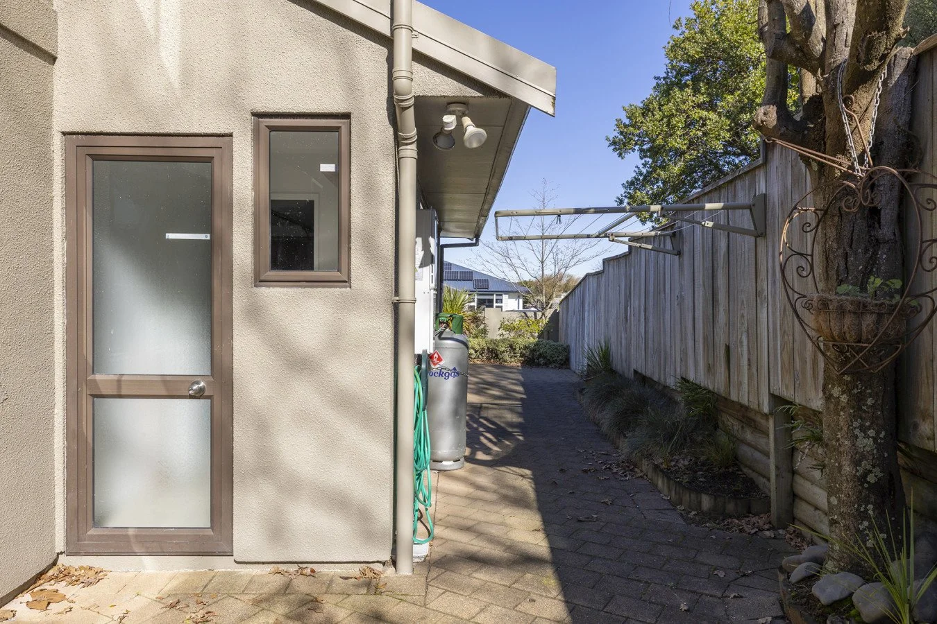 Side yard with a paved pathway, fence on the right, house wall with a door and small window on the left, a propane tank, and a tree with a heart-shaped metal frame around it.