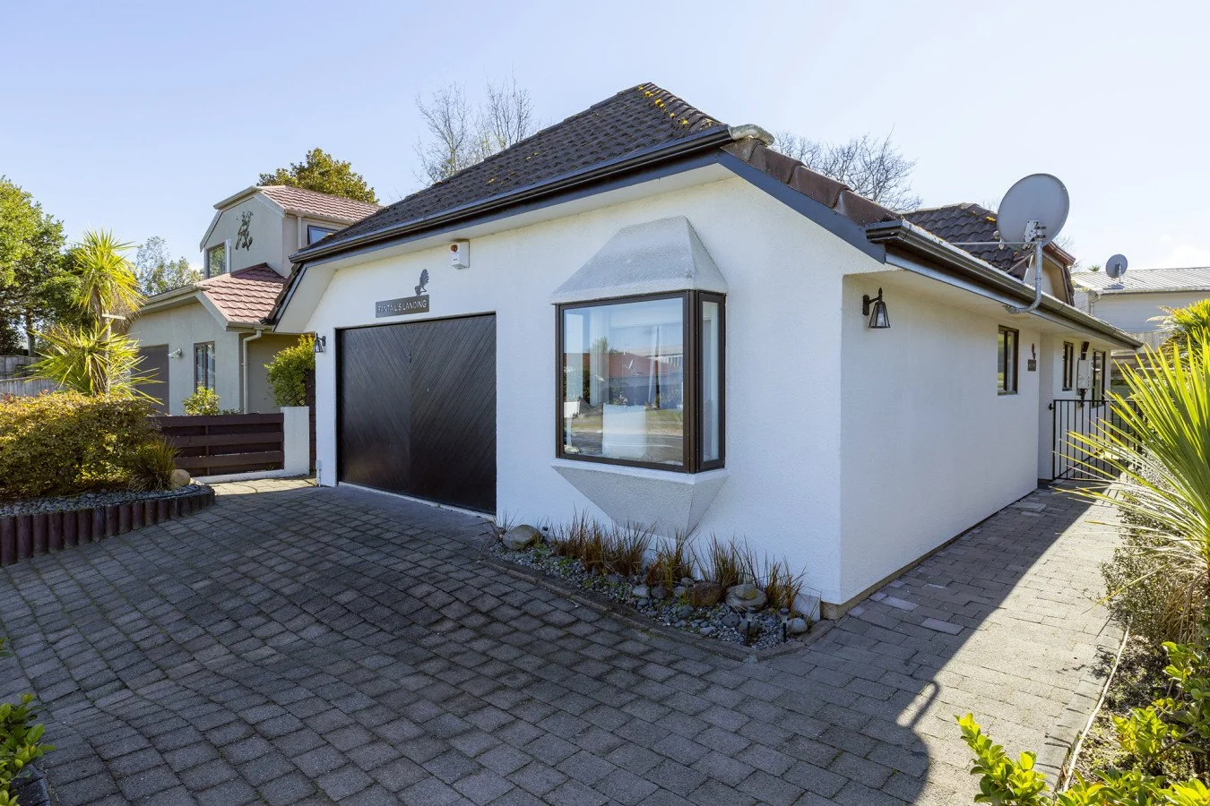 Front view of a white house with a black garage door, large bay window, and satellite dishes on the roof; landscaped driveway and side walkway with plants and bushes.