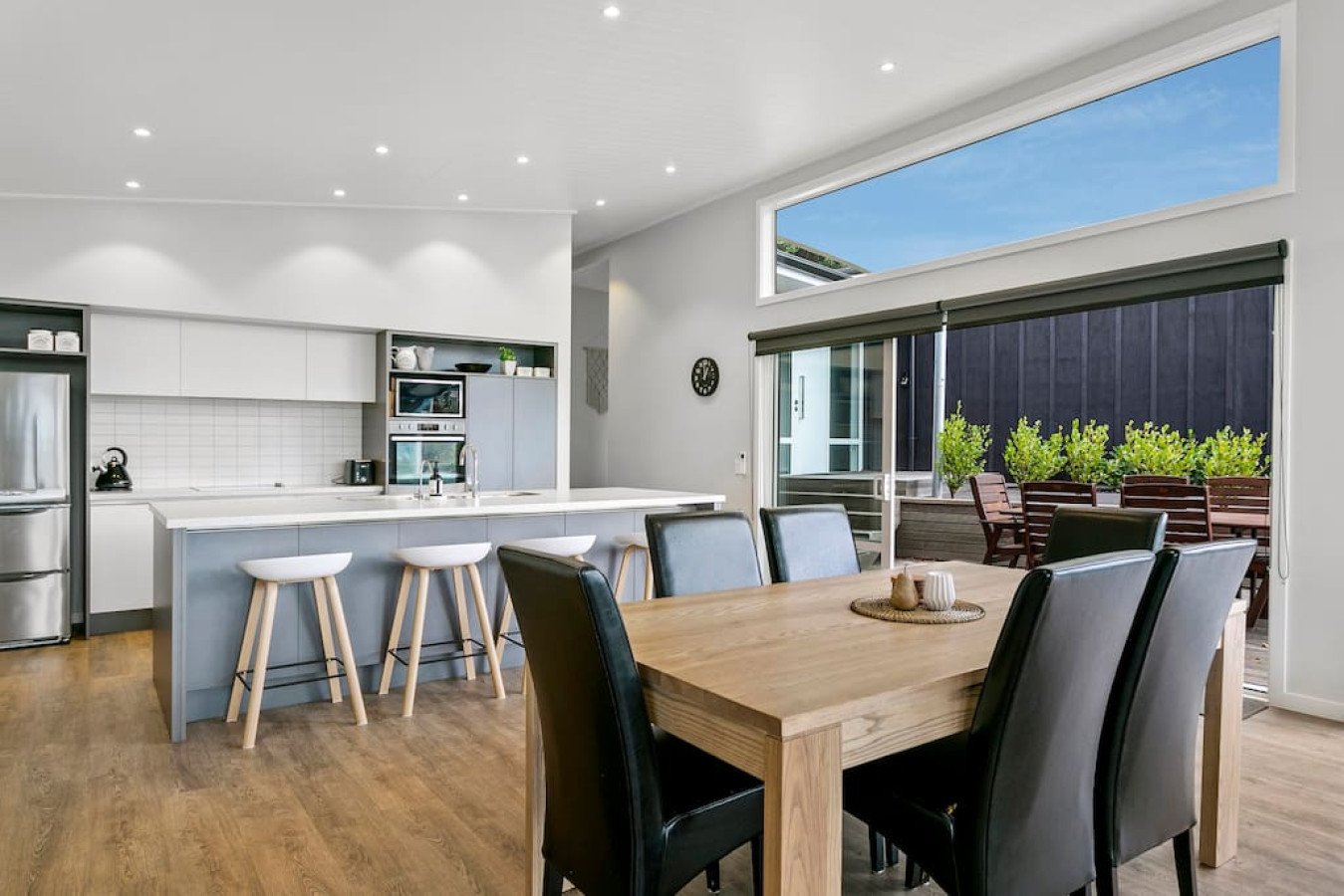 Modern open-plan kitchen and dining area with white cabinets, a wooden dining table with six black chairs, a kitchen island with three white stools, large sliding glass doors leading to an outdoor patio with wooden furniture and greenery, hardwood fl