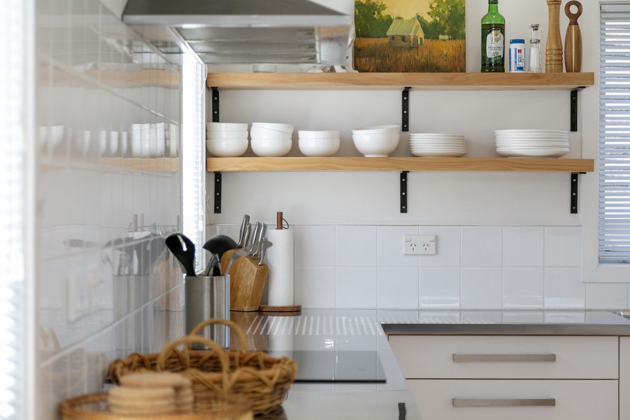 Open kitchen shelf with white bowls and plates, a painting, bottles, and wooden kitchen utensils