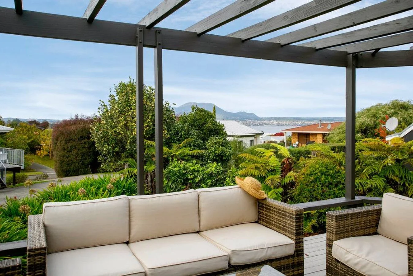 Outdoor patio with beige wicker seating and a straw sun hat on the armrest, overlooking a lush green garden with trees and bushes, with a view of houses, a body of water, and mountains in the distance under a partly cloudy sky.