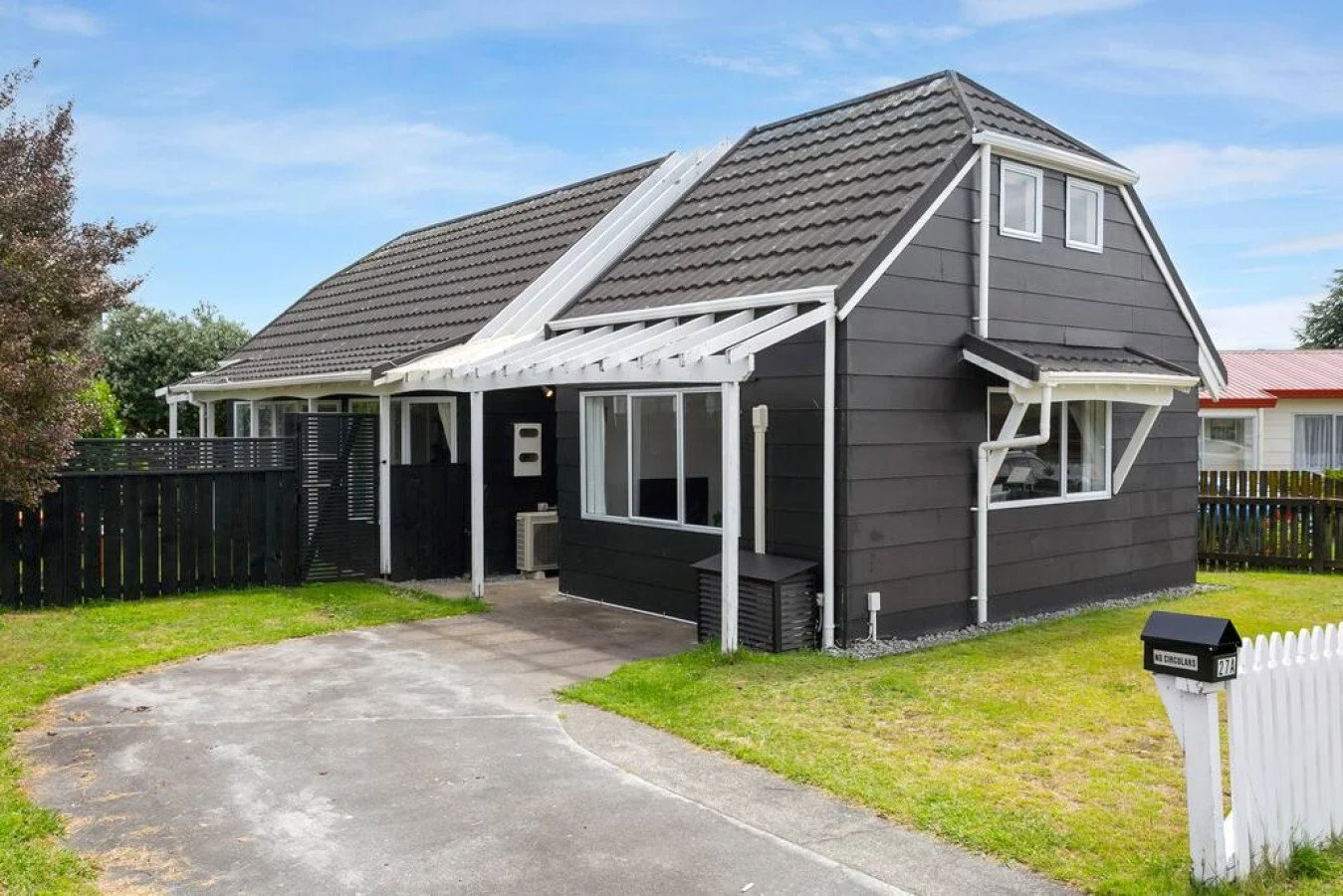A black house with white trim in a suburban neighborhood with a black picket fence, driveway, and lawn.