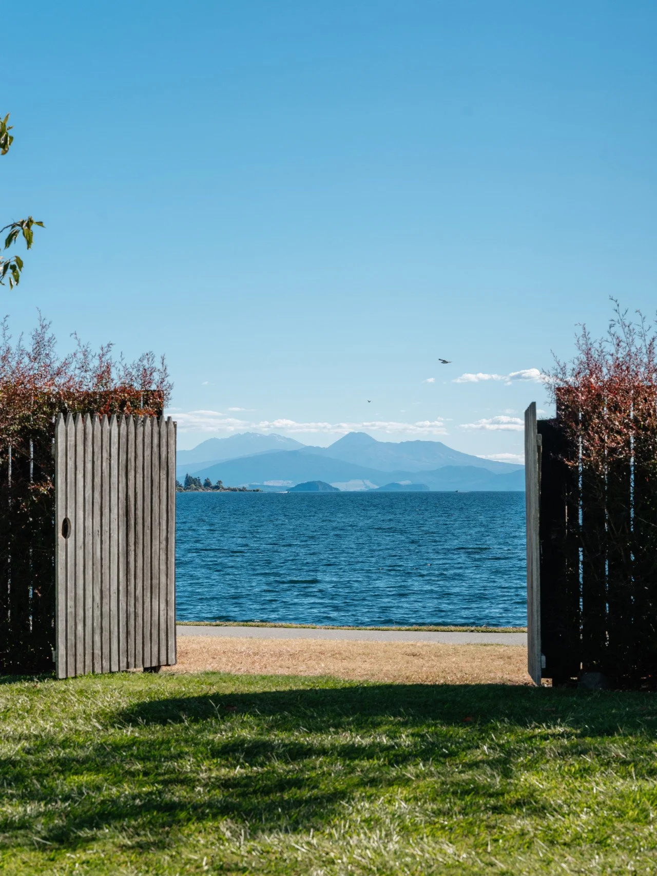 View of a lake with mountains in the distance, seen through an open wooden gate, with greenery and bushes surrounding the area.