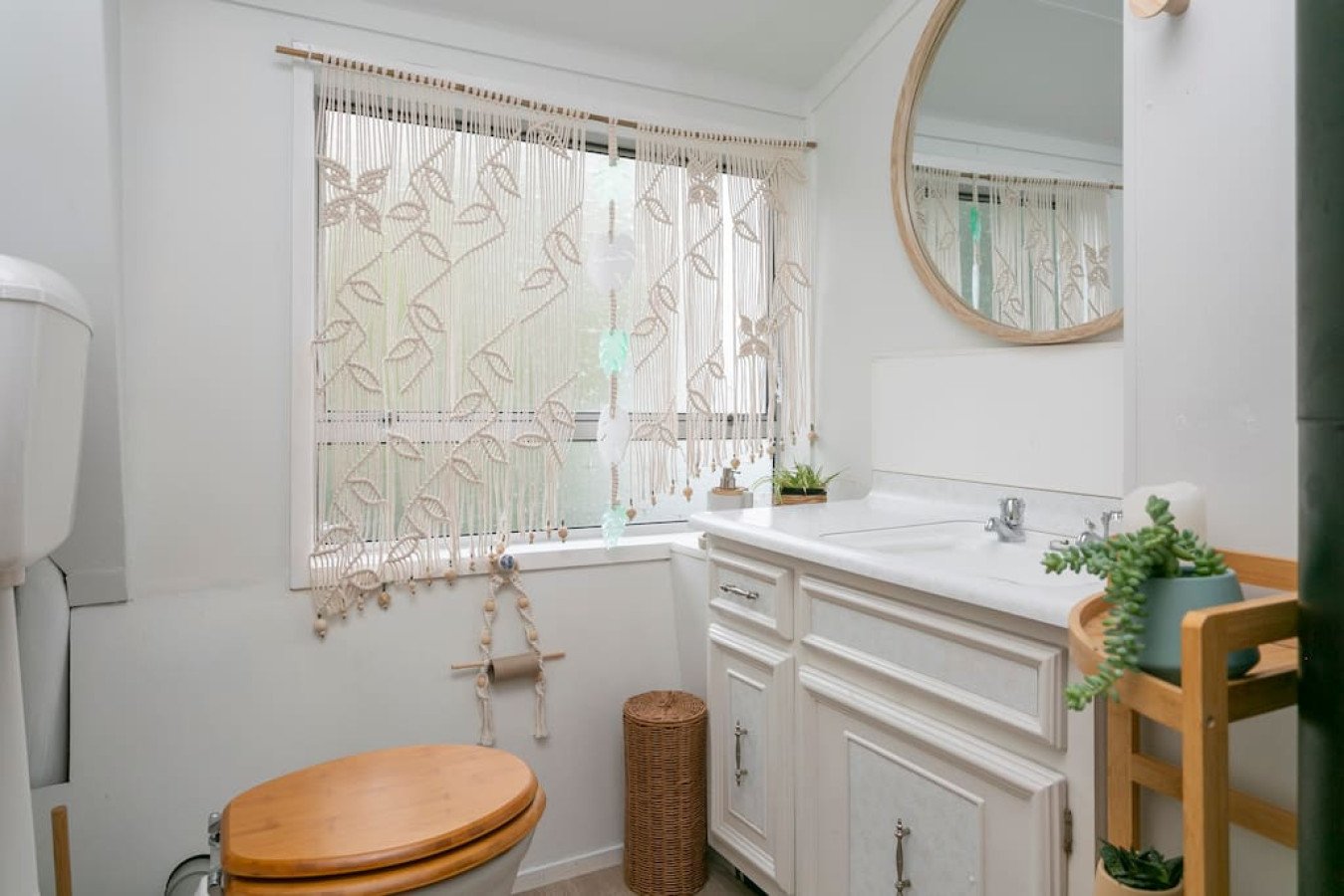 A small bright bathroom with a window covered by decorative curtains, a white vanity with a mirror, a wicker laundry basket, a potted plant, and a toilet with a wooden seat.
