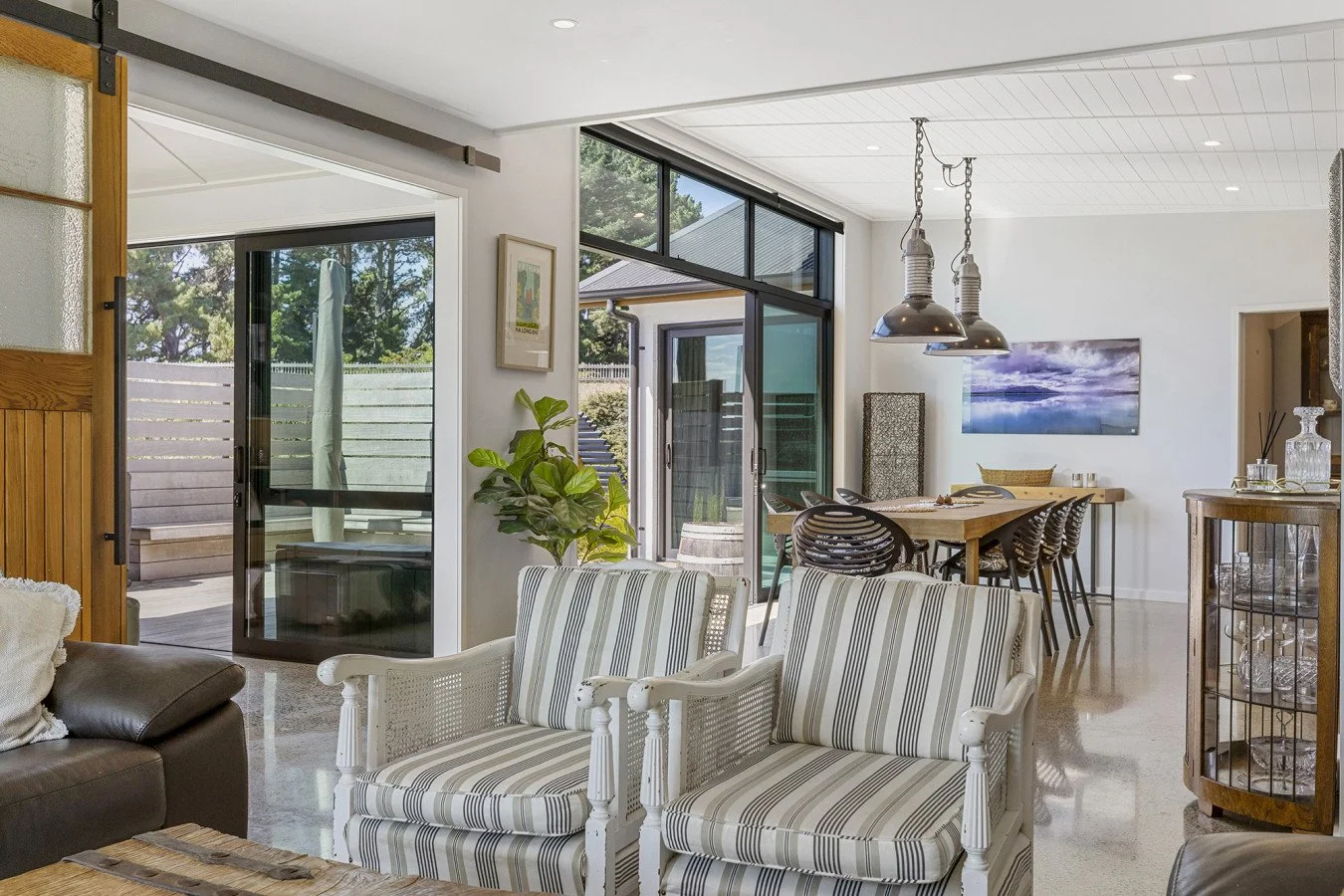 Bright open-concept living and dining area with white walls, glass sliding doors, and wooden furniture, featuring striped armchairs, a dining table with chairs, a large wall art, and natural light.